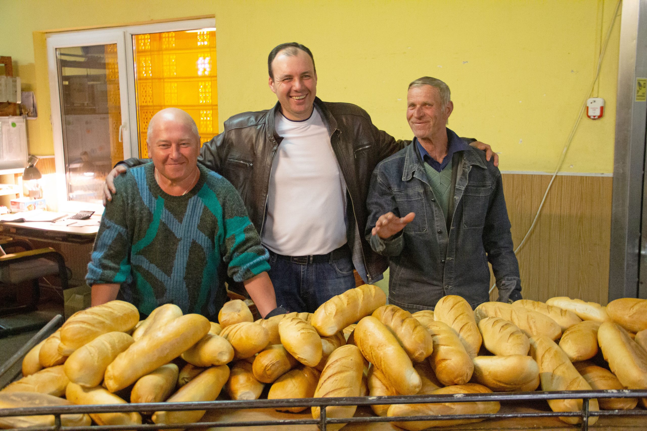 Three men smile in front of a table of bread.